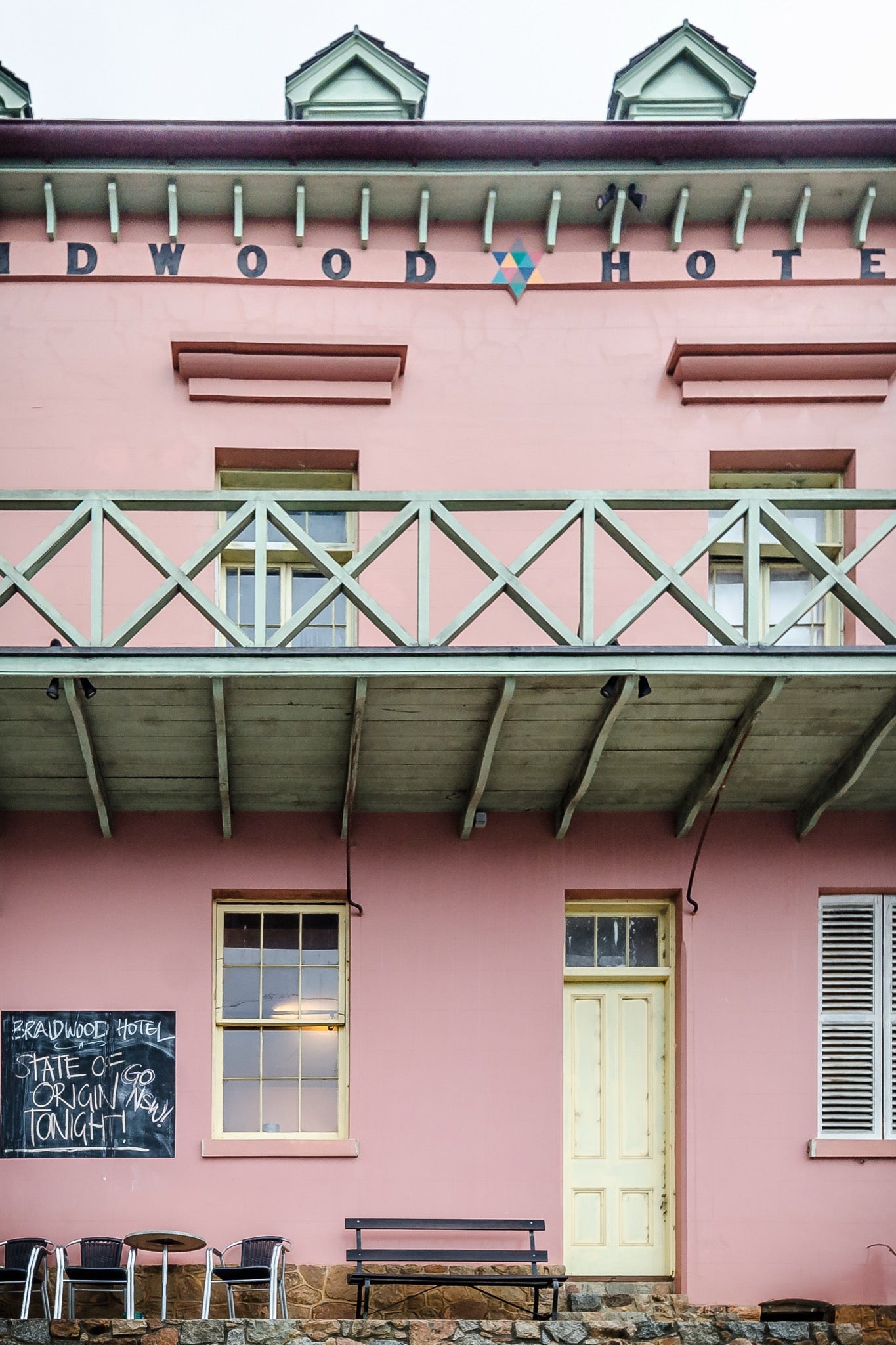 The pink rendered two-storey facade of Braidwood Hotel, with a timber balcony above a verandah, a chalkboard sign, and outdoor seating at street level.