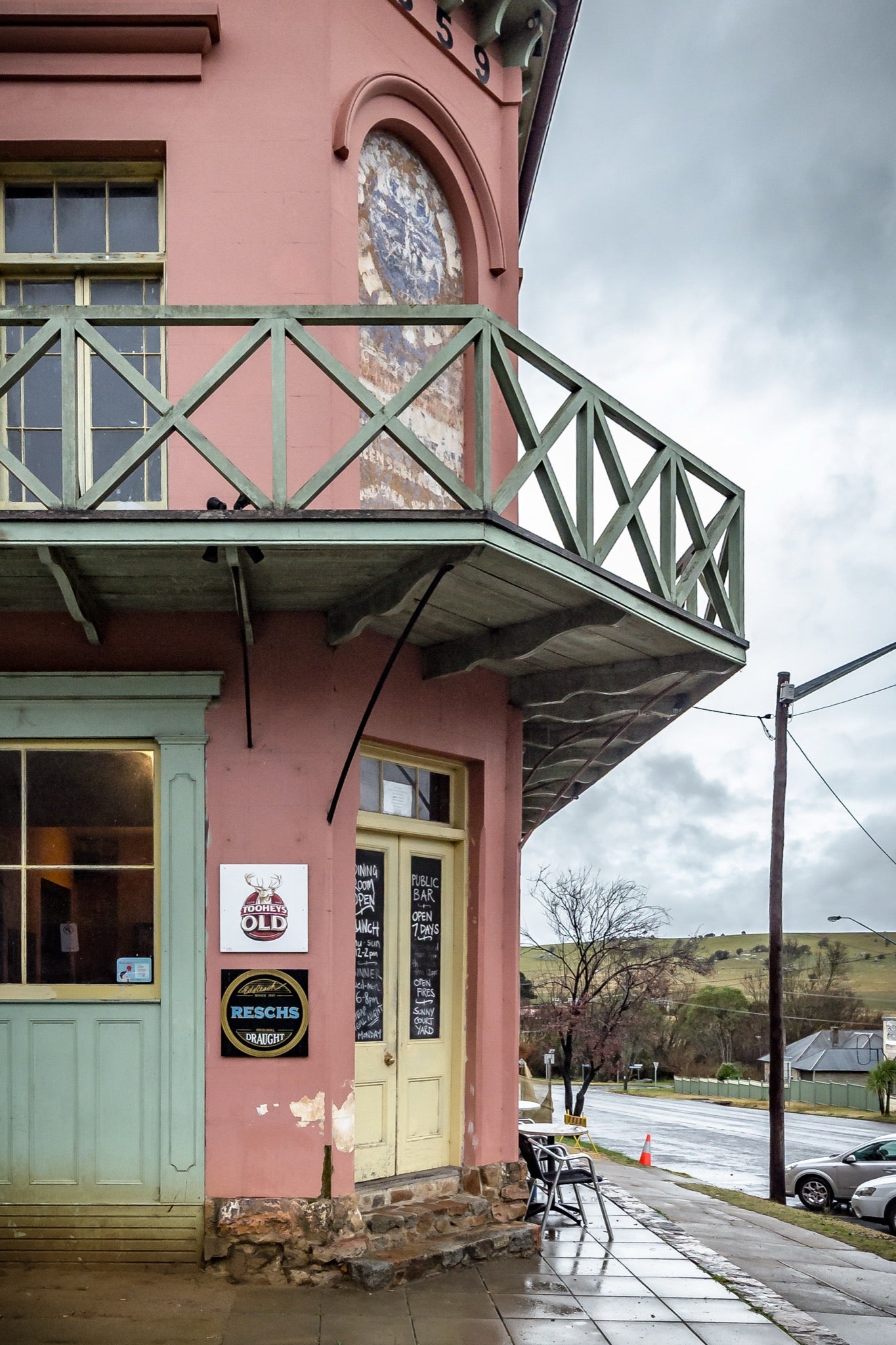 Pink rendered two-storey hotel on a corner footpath at Braidwood, with green timber balcony railings, yellow bar doors, beer signage, and metal tables on wet pavement under an overcast sky.