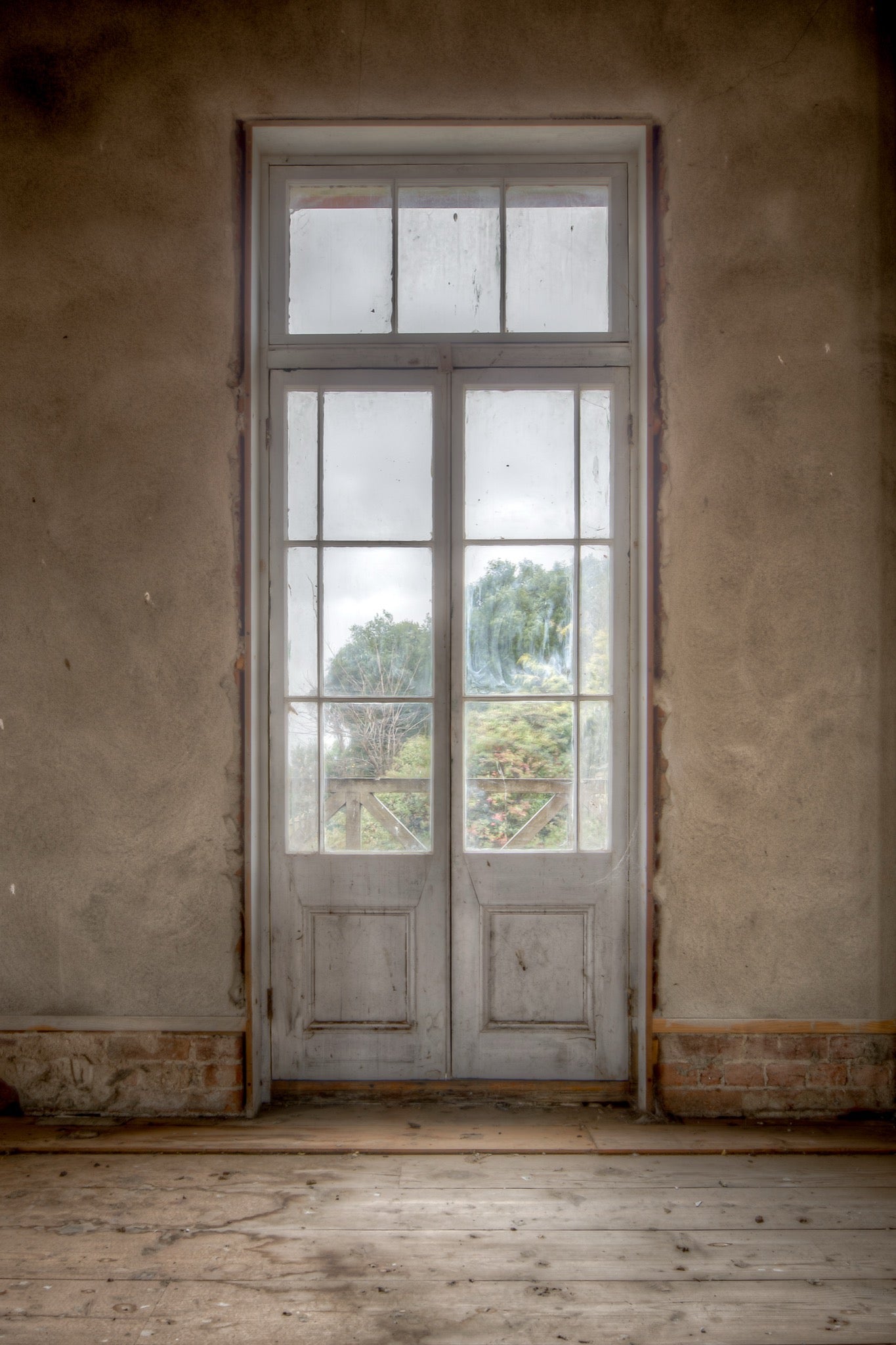 Dusty white-framed French doors with multiple glass panes and timber lower panels, set into a worn rendered wall at the Braidwood Hotel, with green trees visible beyond a timber railing.
