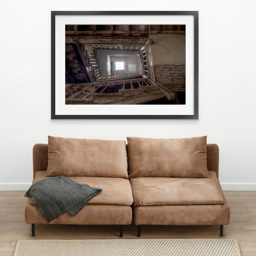 Looking up through the timber stairwell at the Braidwood Hotel, with joists, beams and stair stringers framing a central void rising to a bright window, and cracked rendered walls showing exposed lath behind missing plaster.
