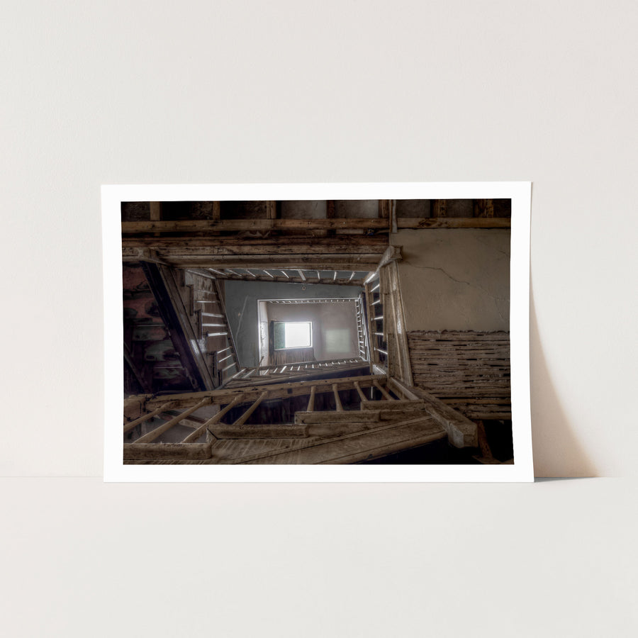 Looking up through the timber stairwell at the Braidwood Hotel, with joists, beams and stair stringers framing a central void rising to a bright window, and cracked rendered walls showing exposed lath behind missing plaster.