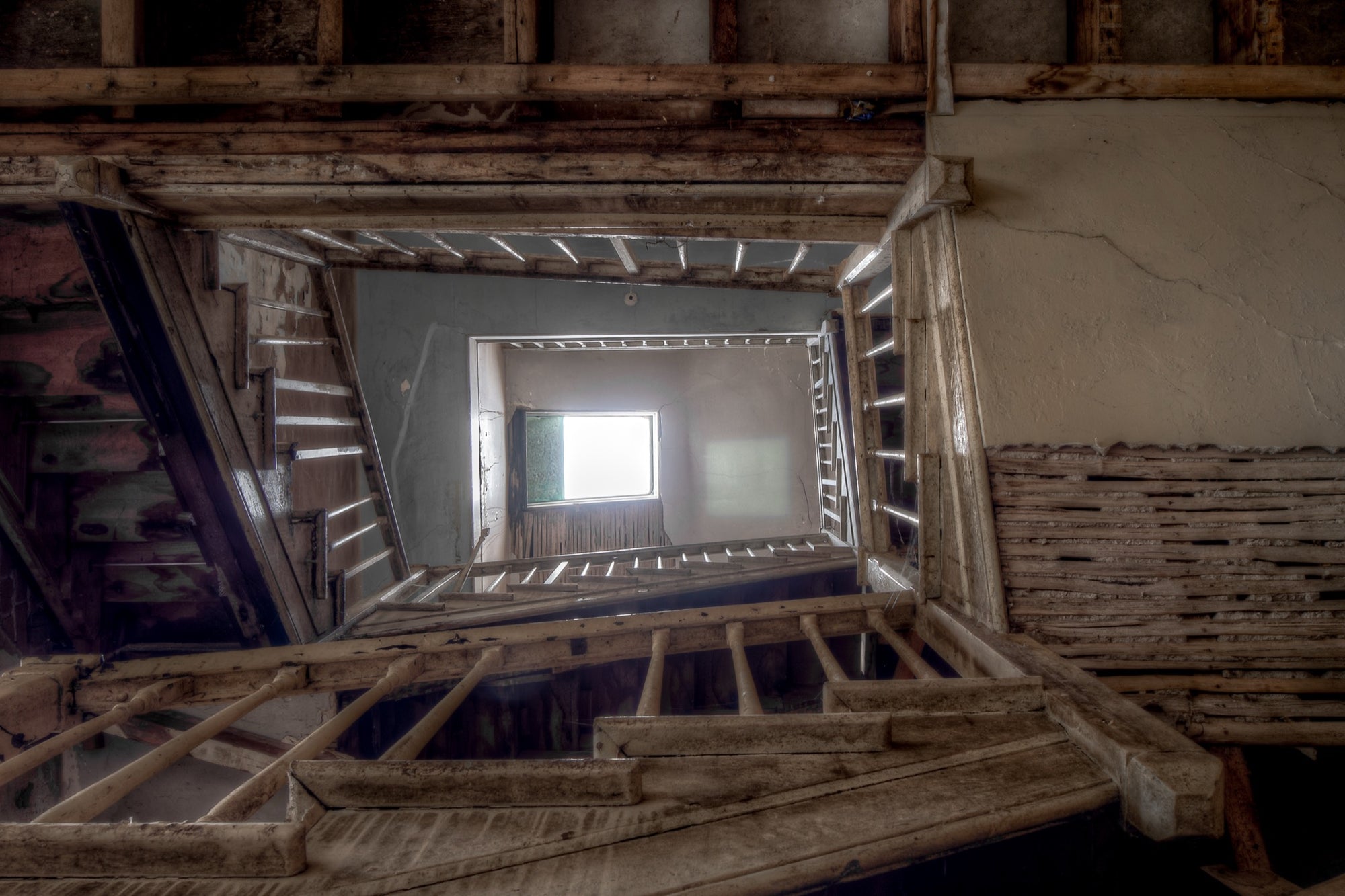 Looking up through the timber stairwell at the Braidwood Hotel, with joists, beams and stair stringers framing a central void rising to a bright window, and cracked rendered walls showing exposed lath behind missing plaster.