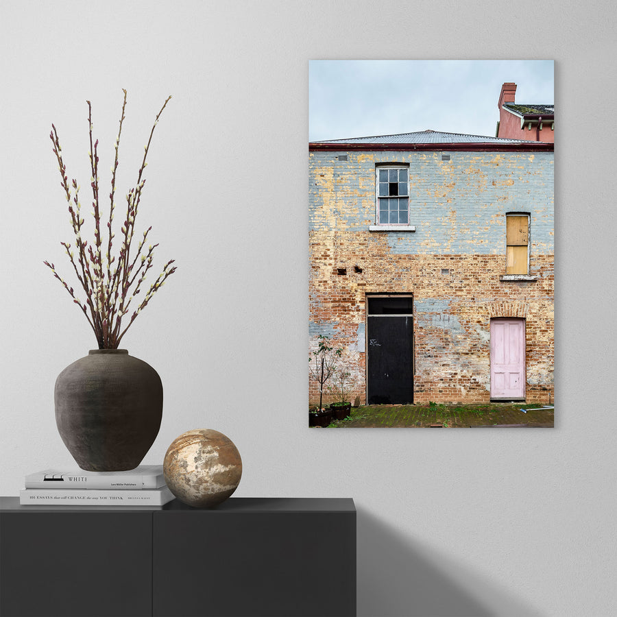 Two ground-level doorways, one black and one pink, set into a weathered rendered brick facade at the Braidwood Hotel, with a six-pane sash window above and a boarded opening to the right beneath corrugated metal roofing.