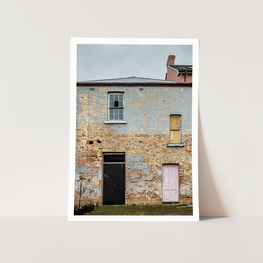 Two ground-level doorways, one black and one pink, set into a weathered rendered brick facade at the Braidwood Hotel, with a six-pane sash window above and a boarded opening to the right beneath corrugated metal roofing.