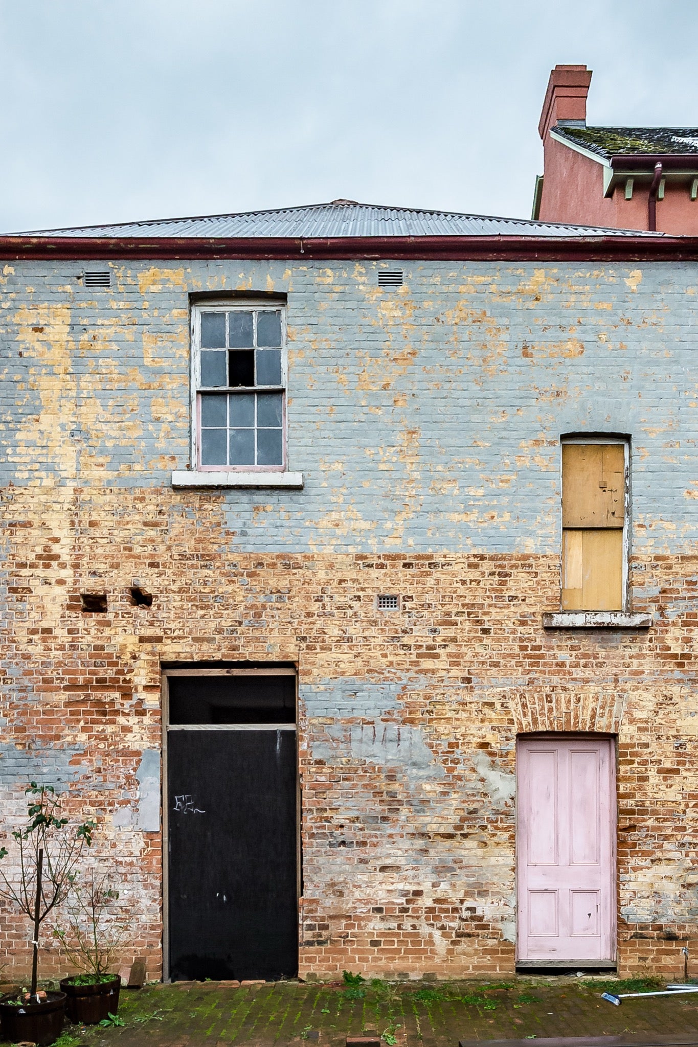 Two ground-level doorways, one black and one pink, set into a weathered rendered brick facade at the Braidwood Hotel, with a six-pane sash window above and a boarded opening to the right beneath corrugated metal roofing.
