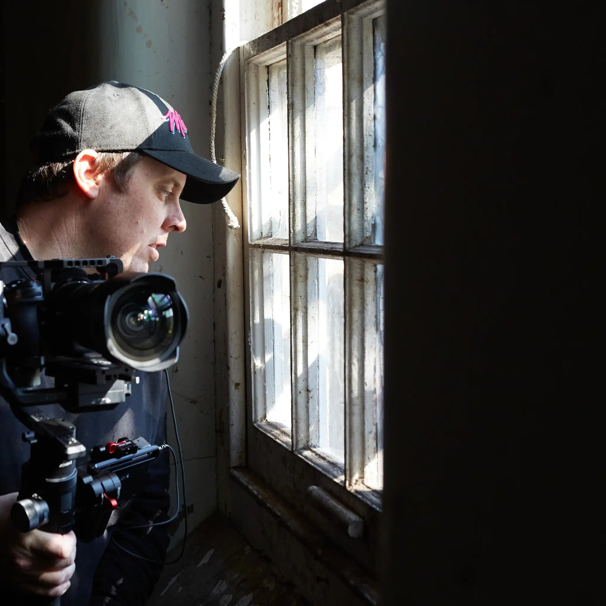 Brett Patman holding a camera inside a dark abandoned building, looking out a bright window, capturing the essence of forgotten spaces.