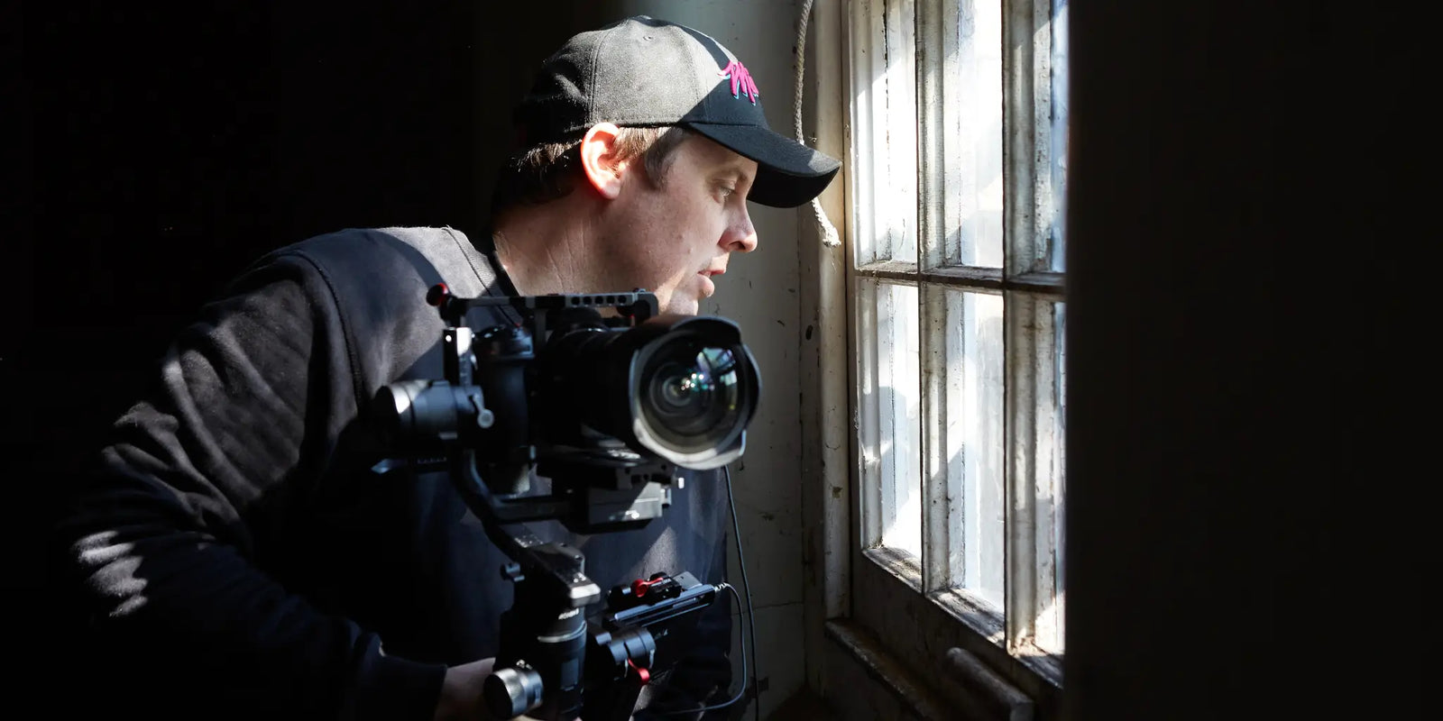 Brett Patman holding a camera inside a dark abandoned building, looking out a bright window, capturing the essence of forgotten spaces.