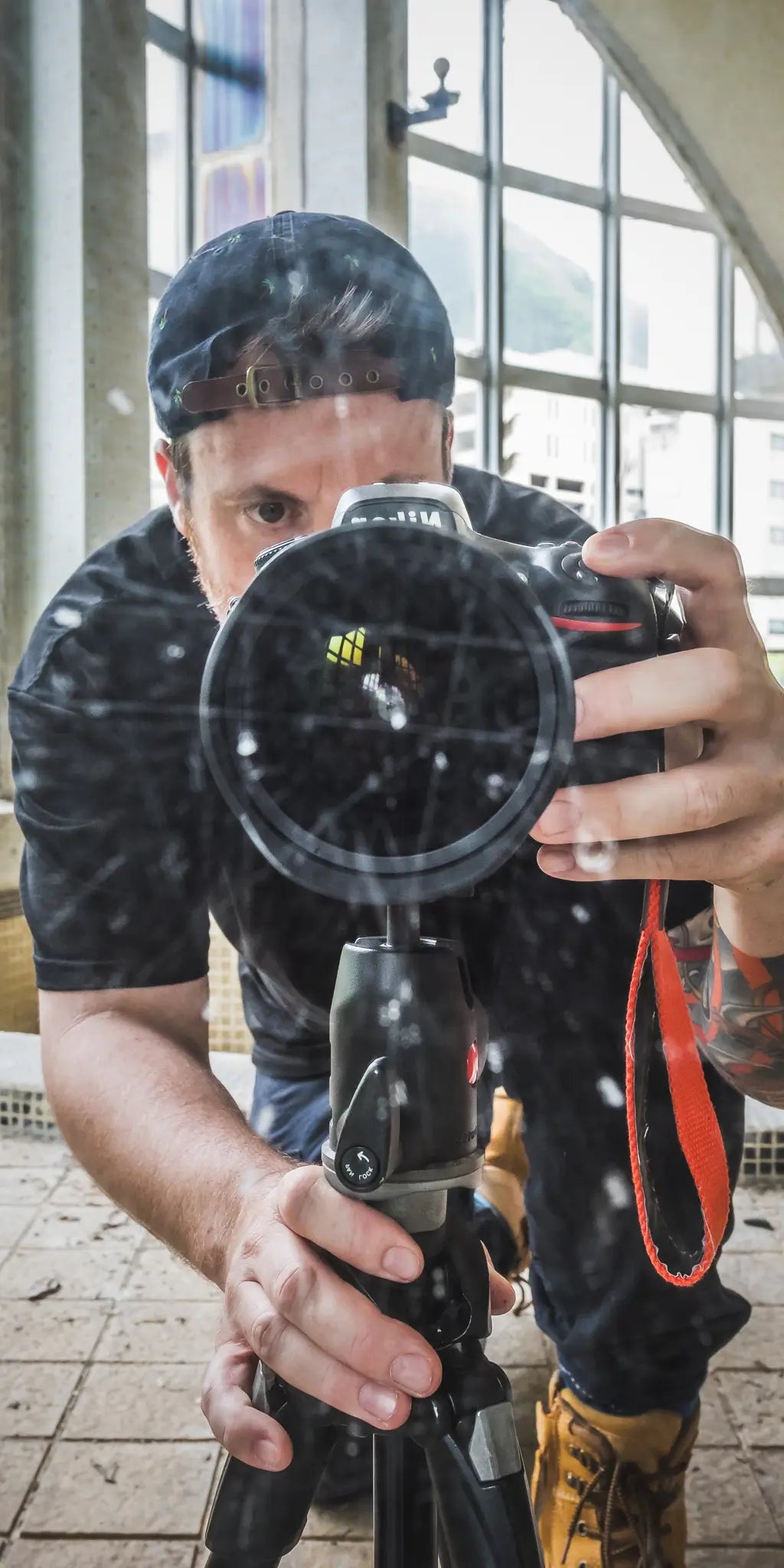 Brett Patman capturing a self-portrait through reflective glass inside an abandoned building, featuring a camera and tripod setup in a naturally lit derelict interior.