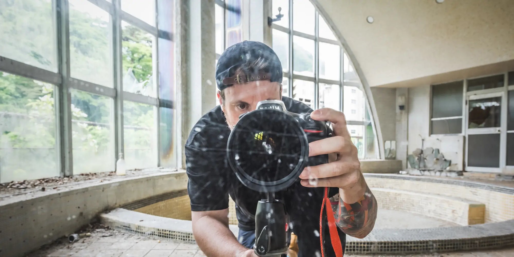 Brett Patman capturing a self-portrait through reflective glass inside an abandoned building, featuring a camera and tripod setup in a naturally lit derelict interior.