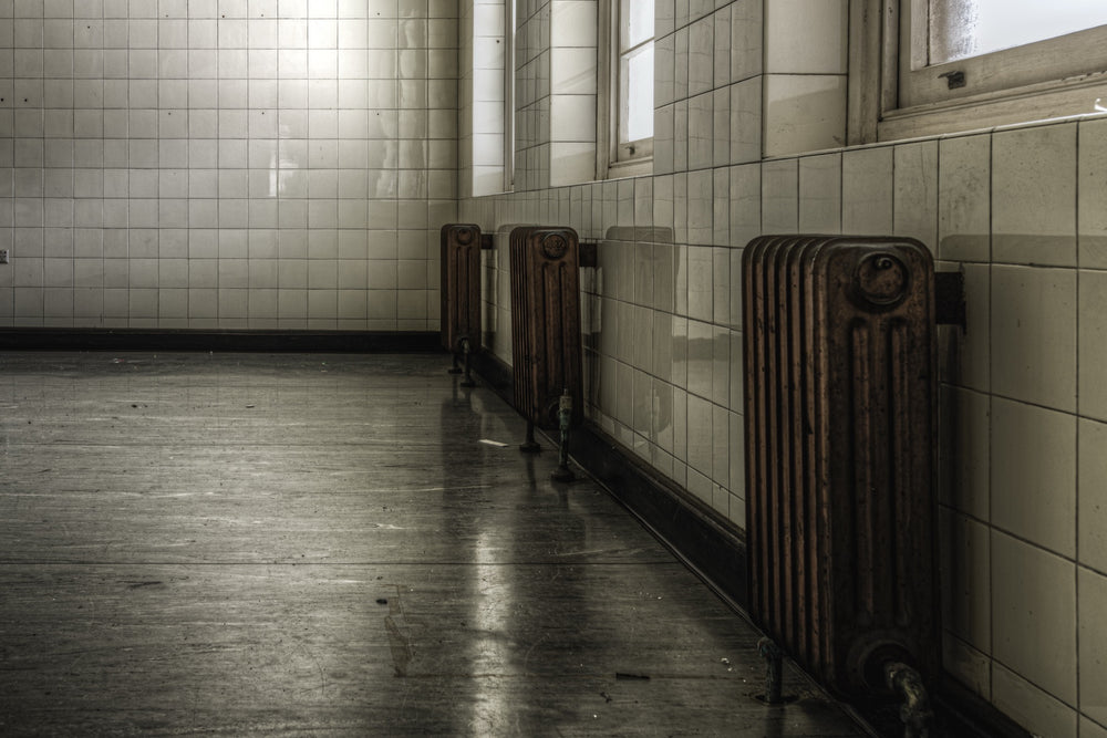 Three cast-iron radiators along a tiled corridor wall at Callan Park, reflected in the polished concrete floor below.