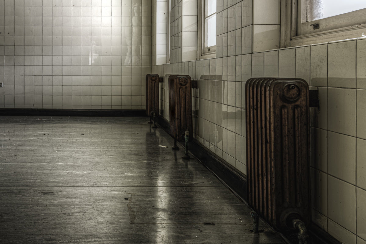 Three cast-iron radiators along a tiled corridor wall at Callan Park, reflected in the polished concrete floor below.