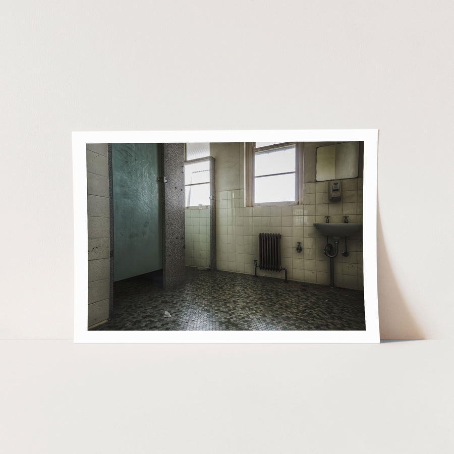 Washroom interior at Callan Park, with lifted hexagonal floor tiles, a white ceramic sink with exposed plumbing, a cast-iron radiator, and light entering through tall timber-framed windows.