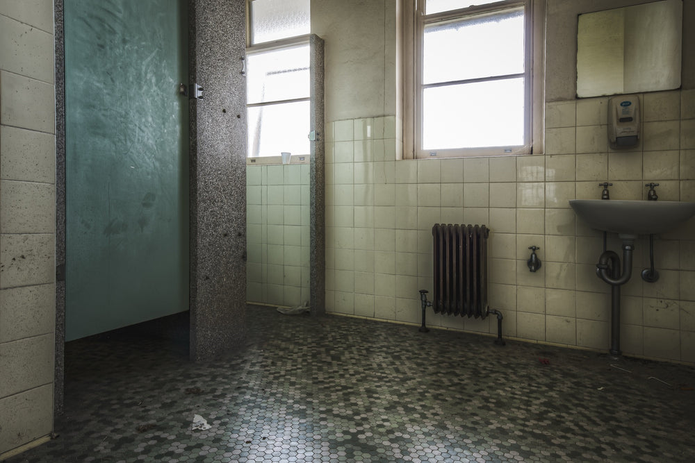Washroom interior at Callan Park, with lifted hexagonal floor tiles, a white ceramic sink with exposed plumbing, a cast-iron radiator, and light entering through tall timber-framed windows.