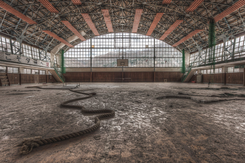 Inside the abandoned gymnasium of Family School Fureai, sunlight illuminates a dusty wooden floor. Faded lines mark former courts.
