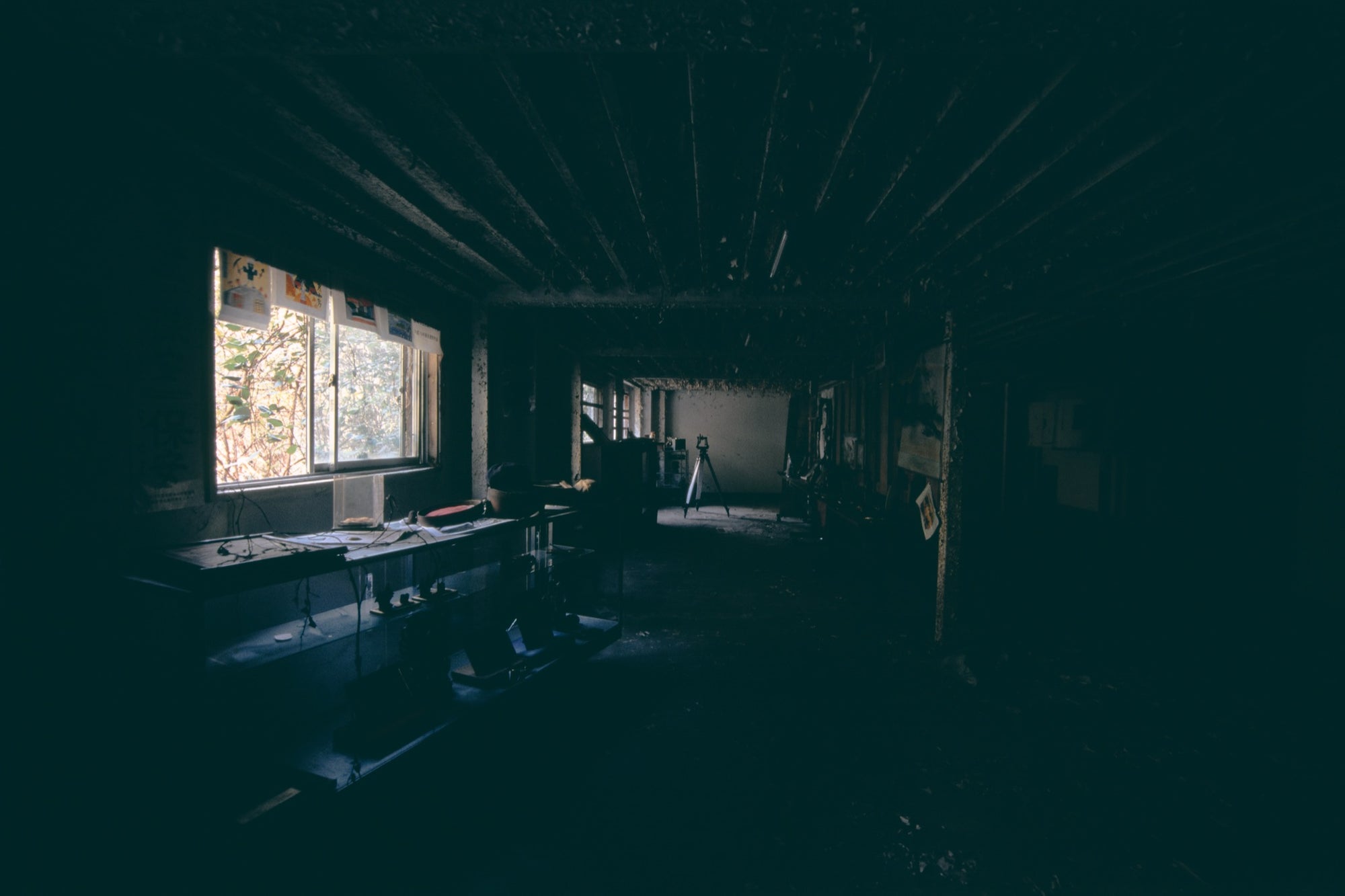 Workbench strewn with wires and objects below three pictures taped to a wall, in the basement of the main mining building at Ikeshima, with foliage at the window and a white tripod lit in the background.