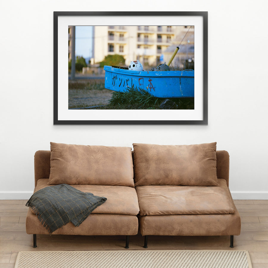 A weathered blue boat with ガンバレ日本 painted on its hull rests on dry grass at Ikeshima, a white ceramic figure inside and a concrete apartment block behind.