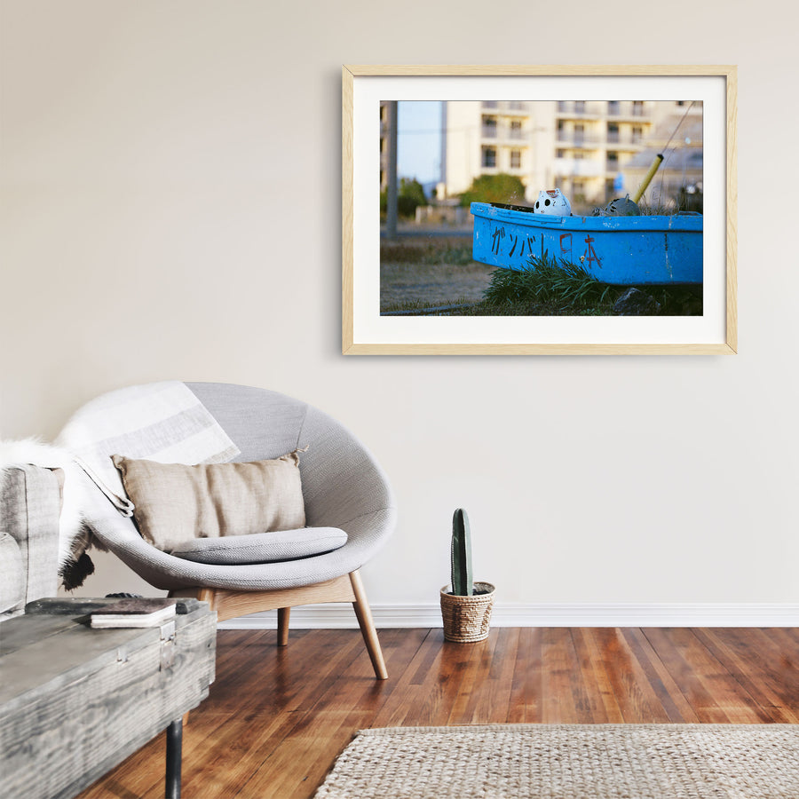 A weathered blue boat with ガンバレ日本 painted on its hull rests on dry grass at Ikeshima, a white ceramic figure inside and a concrete apartment block behind.