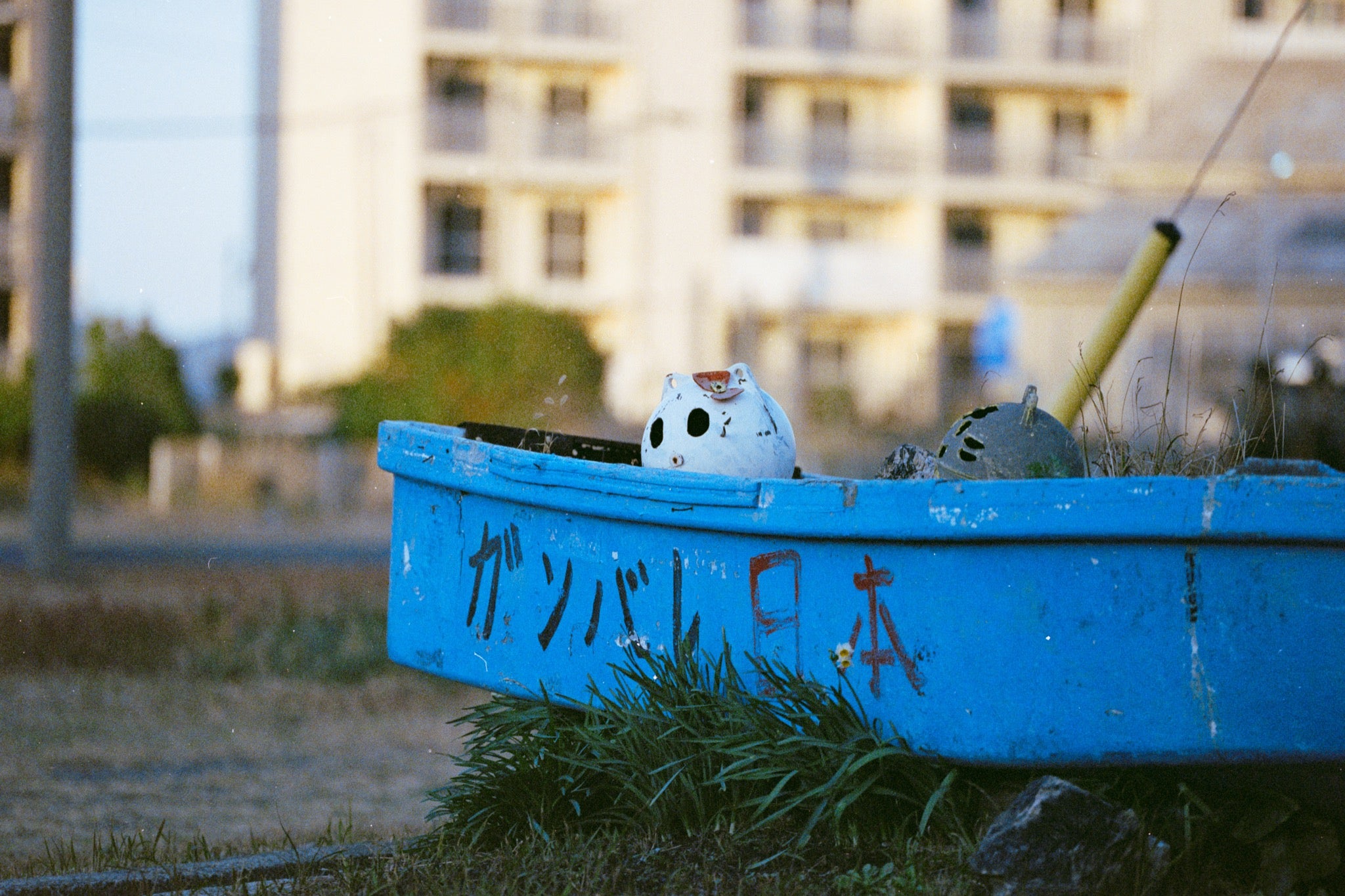 A weathered blue boat with ガンバレ日本 painted on its hull rests on dry grass at Ikeshima, a white ceramic figure inside and a concrete apartment block behind.
