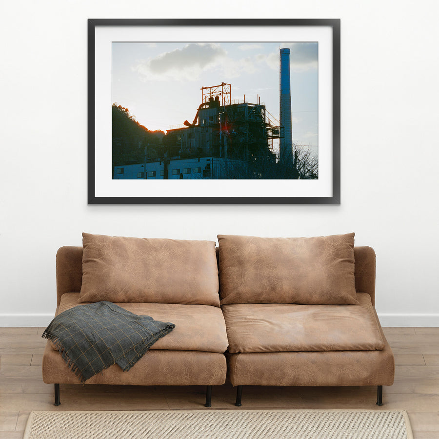 The chimney and boiler house of the coal-fired power station at Ikeshima Coal Mine silhouetted against a hazy sunset sky, with external pipework, steel walkways and a forested hillside visible in the frame.