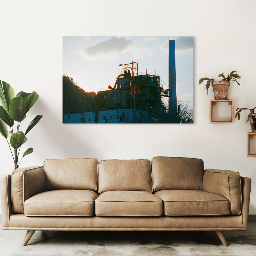 The chimney and boiler house of the coal-fired power station at Ikeshima Coal Mine silhouetted against a hazy sunset sky, with external pipework, steel walkways and a forested hillside visible in the frame.