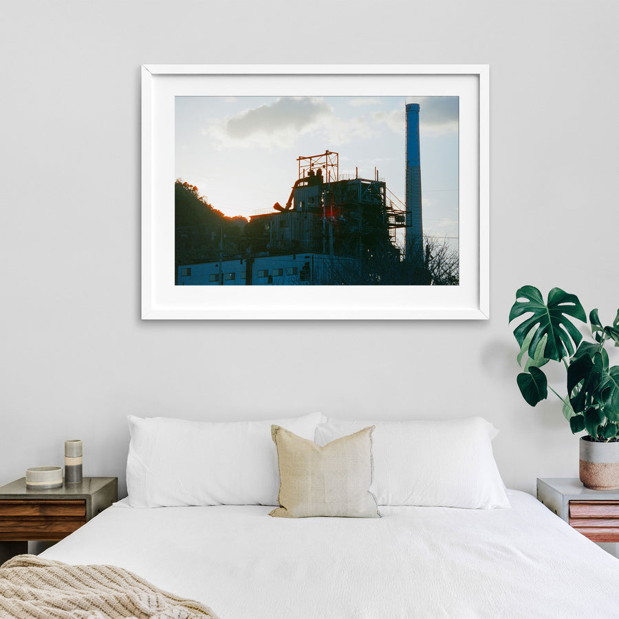The chimney and boiler house of the coal-fired power station at Ikeshima Coal Mine silhouetted against a hazy sunset sky, with external pipework, steel walkways and a forested hillside visible in the frame.