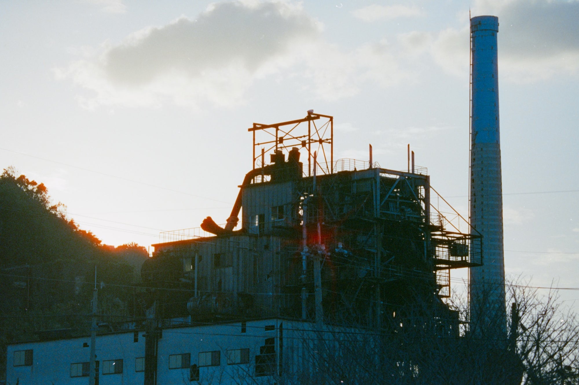 The chimney and boiler house of the coal-fired power station at Ikeshima Coal Mine silhouetted against a hazy sunset sky, with external pipework, steel walkways and a forested hillside visible in the frame.