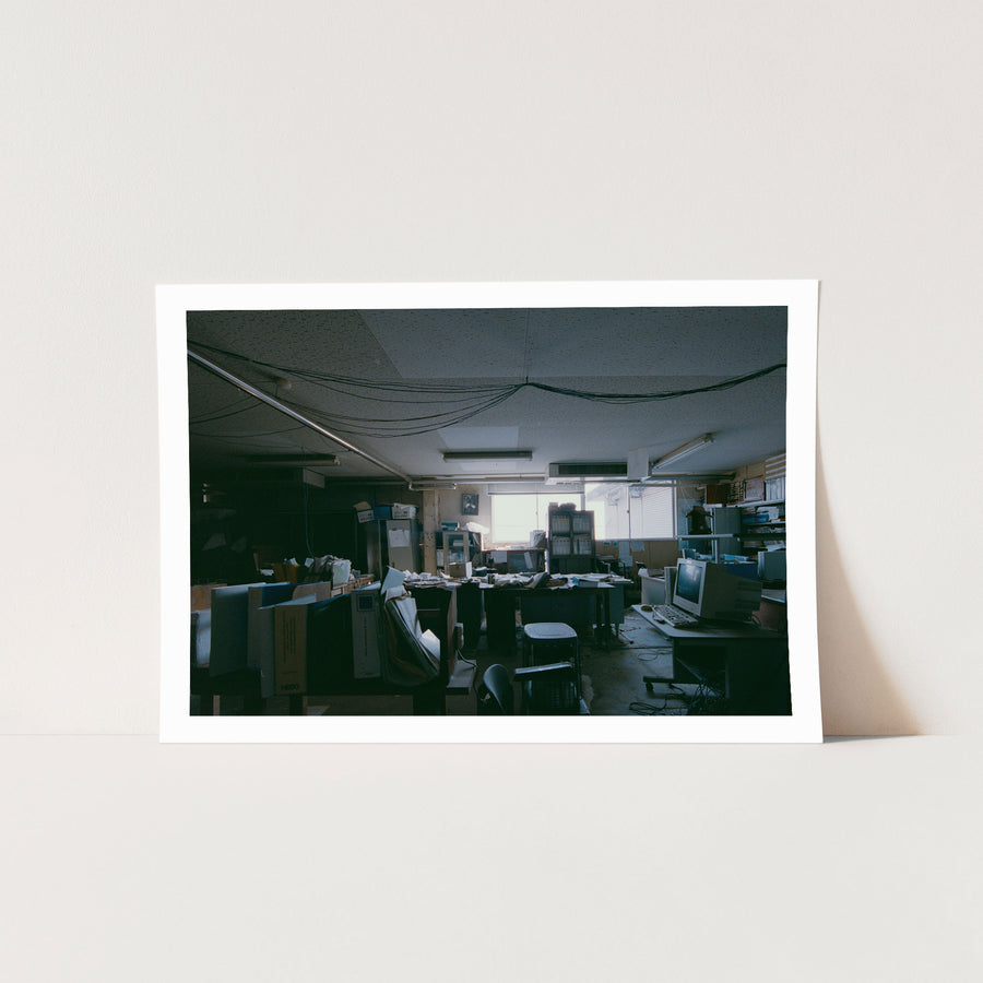 A cluttered desk covered in ring binders, paperwork and a white NEDO box, with a framed flamingo print on the wall above a cabinet, photographed inside the coal mine offices at Ikeshima.