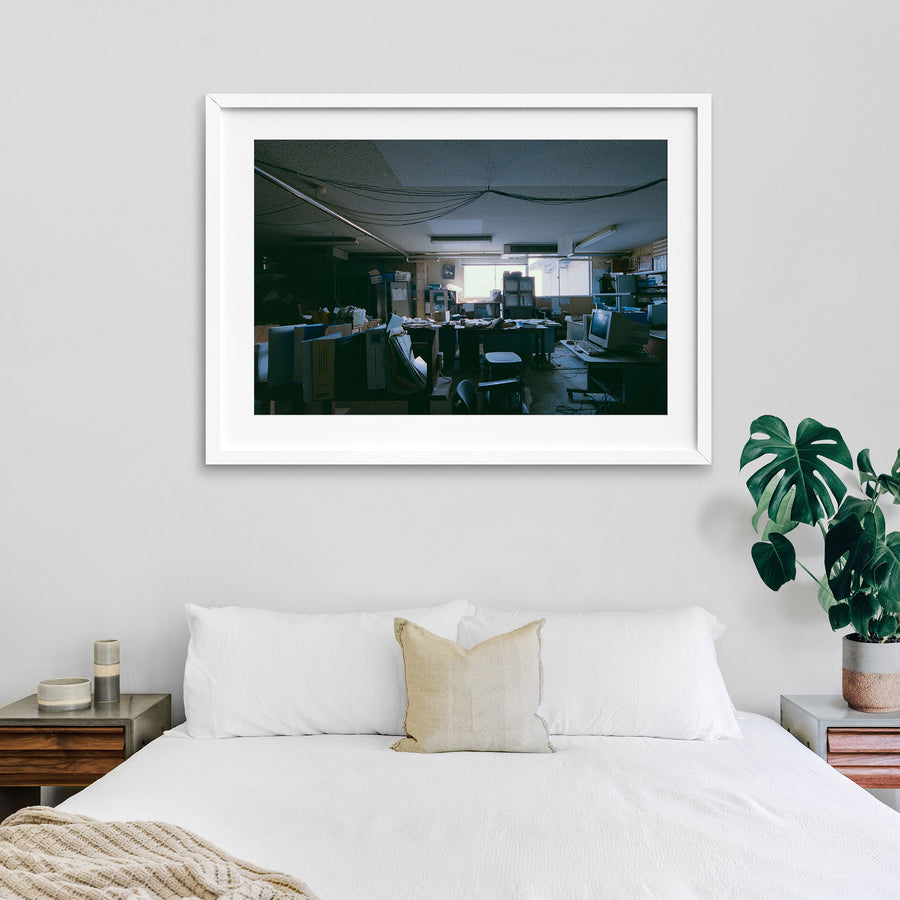 A cluttered desk covered in ring binders, paperwork and a white NEDO box, with a framed flamingo print on the wall above a cabinet, photographed inside the coal mine offices at Ikeshima.