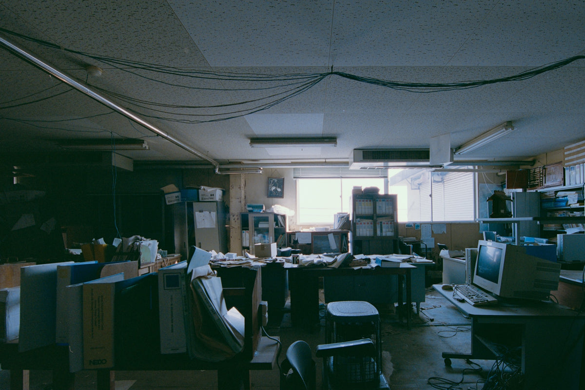 A cluttered desk covered in ring binders, paperwork and a white NEDO box, with a framed flamingo print on the wall above a cabinet, photographed inside the coal mine offices at Ikeshima.