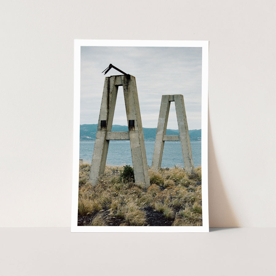 Two concrete A-frame structures with a broken metal arm at the apex of the taller frame, standing on a coastal outcrop at Ikeshima above blue ocean.
