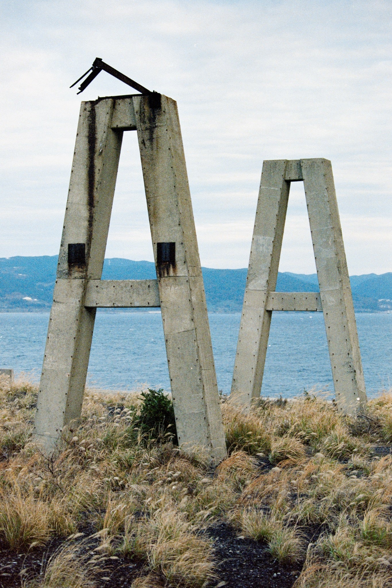 Two concrete A-frame structures with a broken metal arm at the apex of the taller frame, standing on a coastal outcrop at Ikeshima above blue ocean.