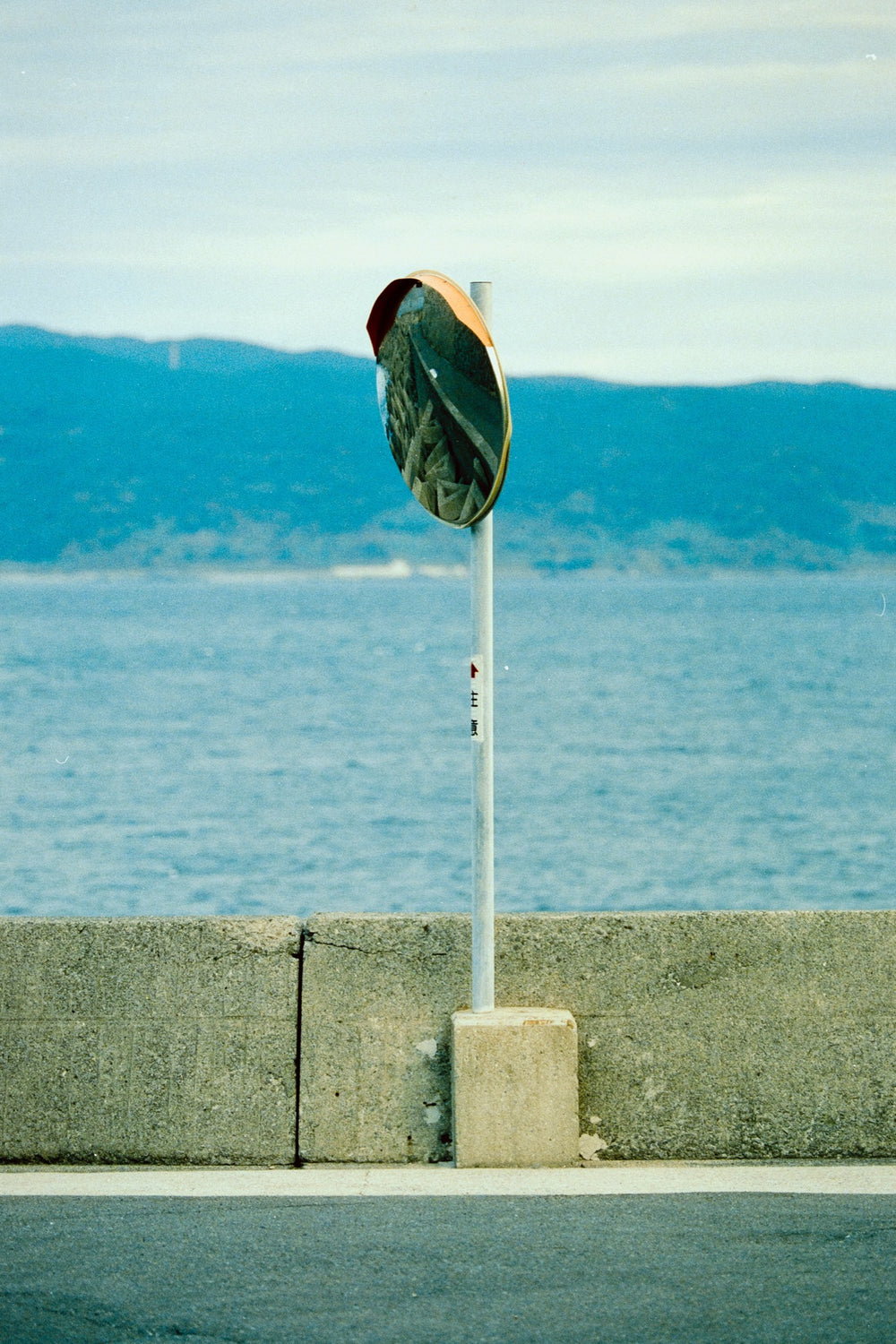 Convex traffic mirror on a white pole at the seawall on Ikeshima, tipped downward with the sea and mainland hills behind.