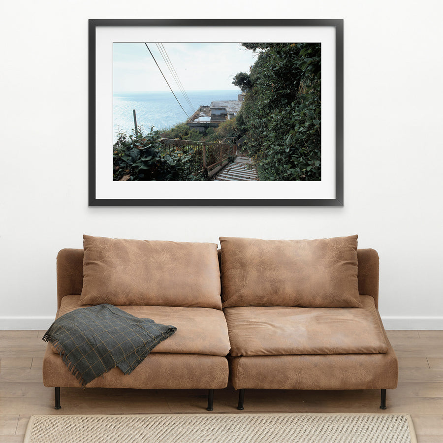 A steep concrete staircase with a rusted railing descends through dense green foliage at Ikeshima, with power lines crossing a bright sky and the sea visible below.