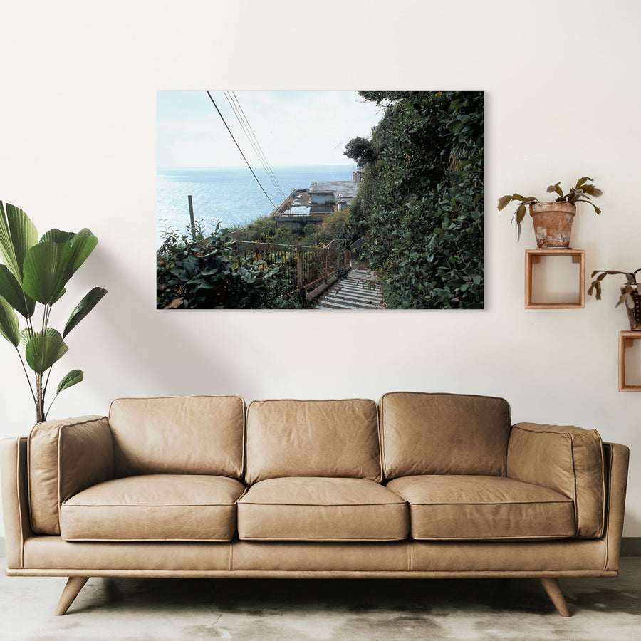 A steep concrete staircase with a rusted railing descends through dense green foliage at Ikeshima, with power lines crossing a bright sky and the sea visible below.