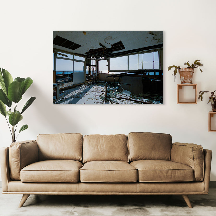 A collapsed ceiling in a disused office at Ikeshima Coal Mine, with a large rectangular opening exposing the cavity above, loose metal grid, plaster debris on the floor, a broken window pane, and a white-topped table near windows that open toward the sea.