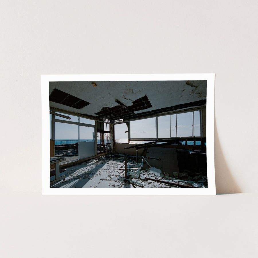A collapsed ceiling in a disused office at Ikeshima Coal Mine, with a large rectangular opening exposing the cavity above, loose metal grid, plaster debris on the floor, a broken window pane, and a white-topped table near windows that open toward the sea.