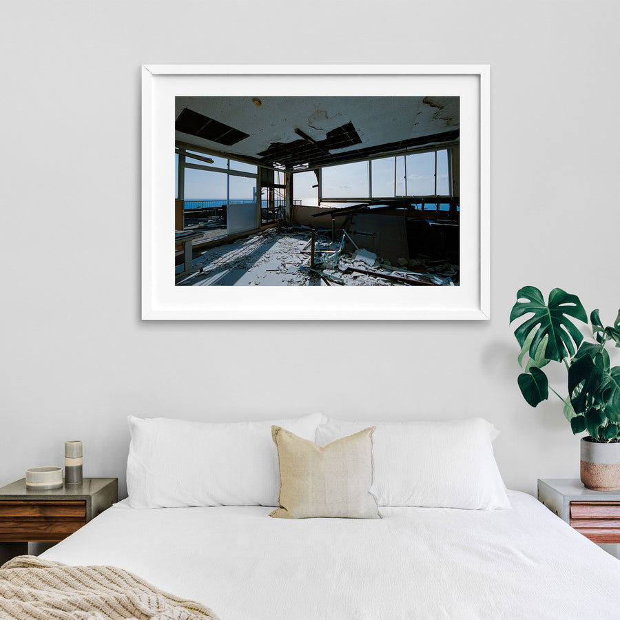 A collapsed ceiling in a disused office at Ikeshima Coal Mine, with a large rectangular opening exposing the cavity above, loose metal grid, plaster debris on the floor, a broken window pane, and a white-topped table near windows that open toward the sea.