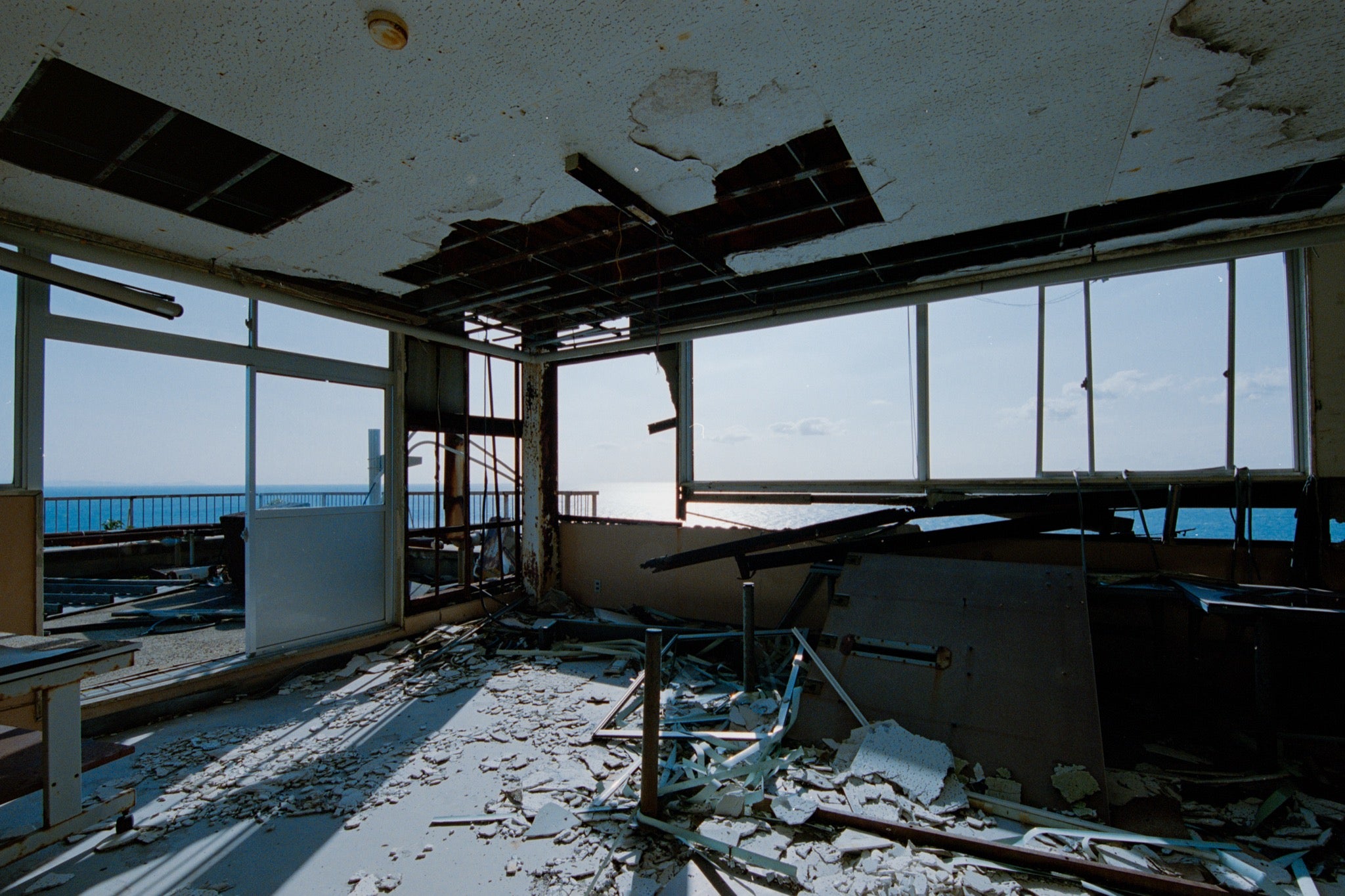 A collapsed ceiling in a disused office at Ikeshima Coal Mine, with a large rectangular opening exposing the cavity above, loose metal grid, plaster debris on the floor, a broken window pane, and a white-topped table near windows that open toward the sea.