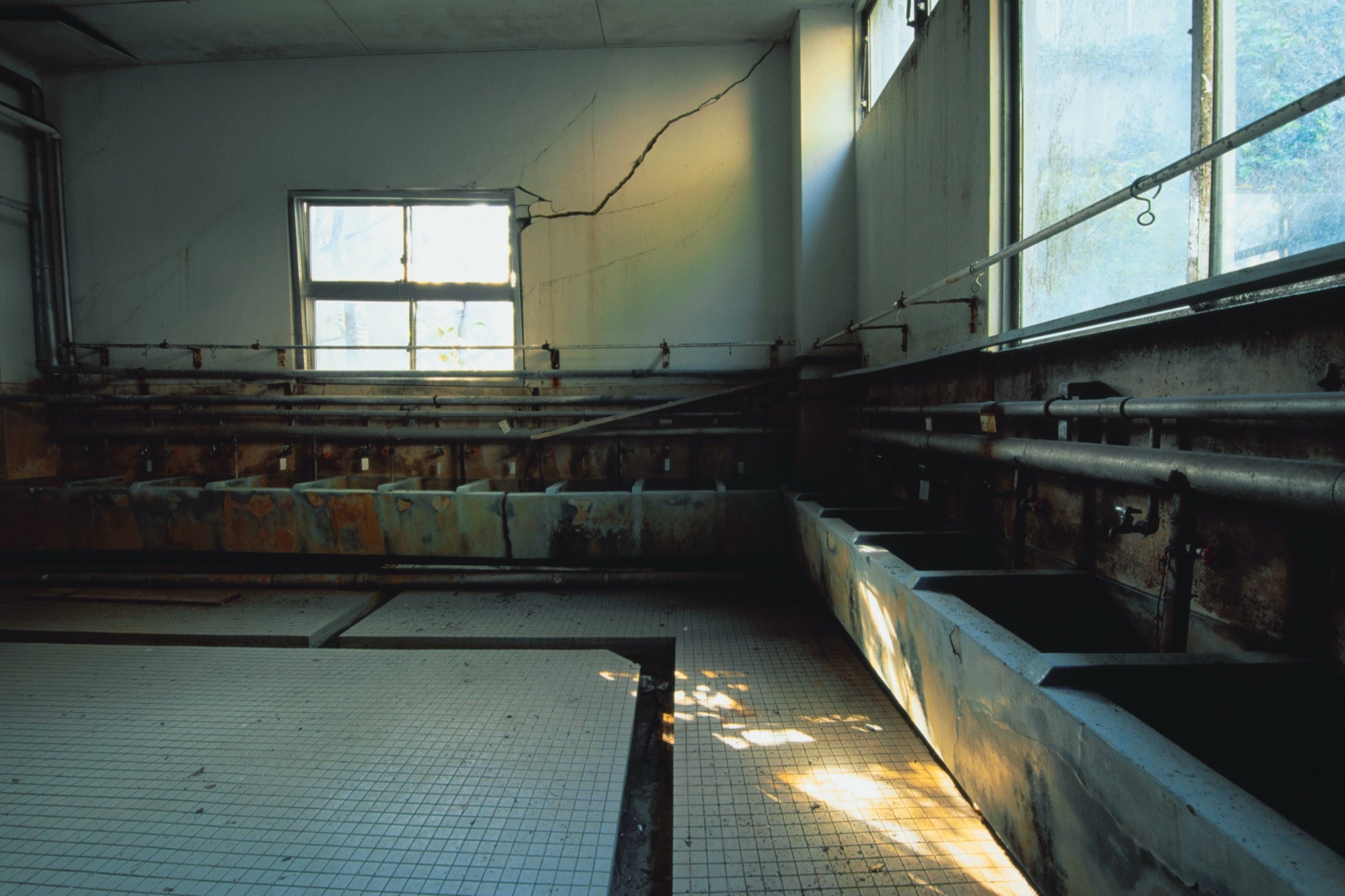 Rusted concrete wash basins and a cracked rear wall inside an abandoned communal bathhouse at Ikeshima, with hard window light crossing the tiled floor.