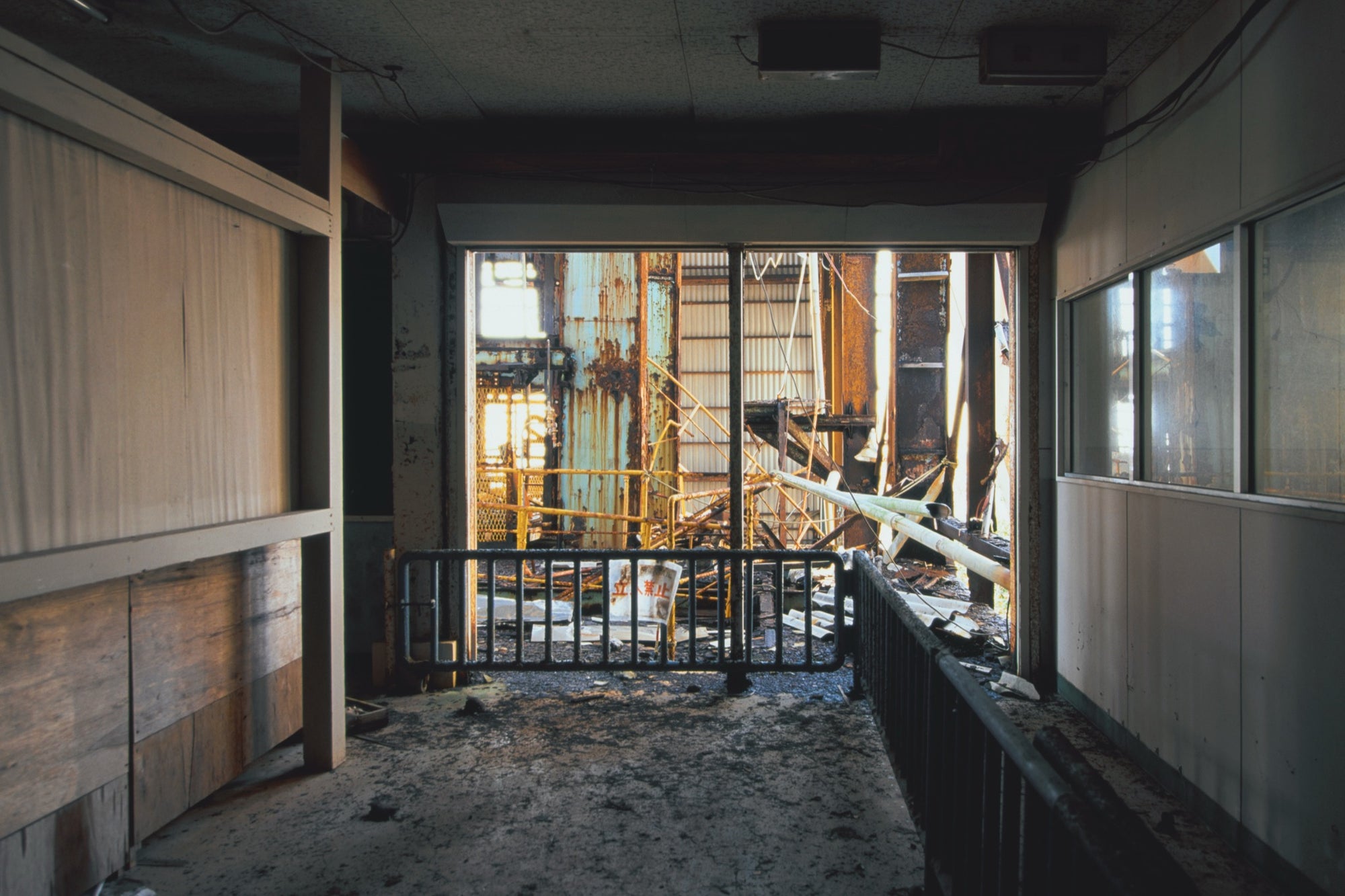 A rusted metal barrier carrying a no-entry sign blocks a doorway in a weathered interior at Ikeshima, with collapsed steelwork and broken pipes visible beyond in bright sunlight.
