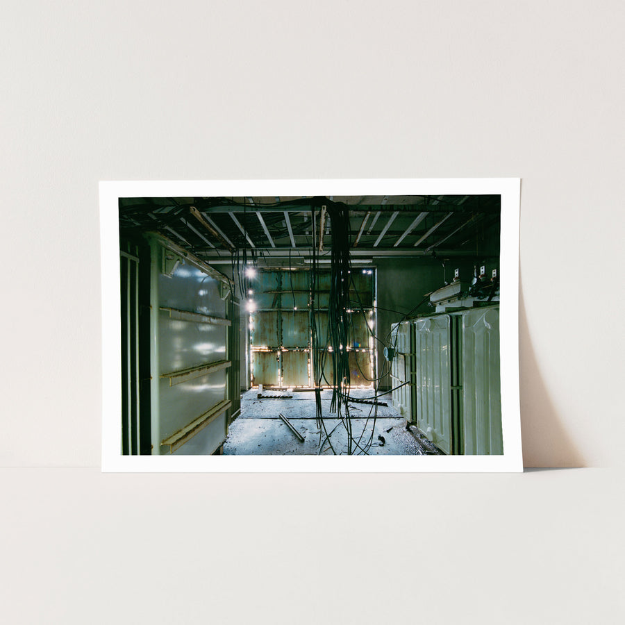 Dense black cable bundle hanging from an exposed ceiling frame runs the length of a corridor at Ikeshima Coal Mine, with two ribbed transformers along one wall and teal double doors at the far end.