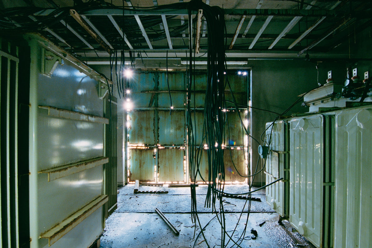 Dense black cable bundle hanging from an exposed ceiling frame runs the length of a corridor at Ikeshima Coal Mine, with two ribbed transformers along one wall and teal double doors at the far end.