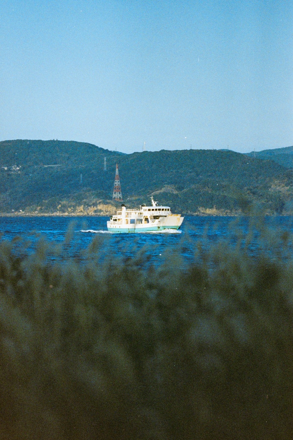 A white and teal ferry crosses the channel at Ikeshima, trailing a bright wake, with a red and white lattice tower rising above the forested hillside behind the port.