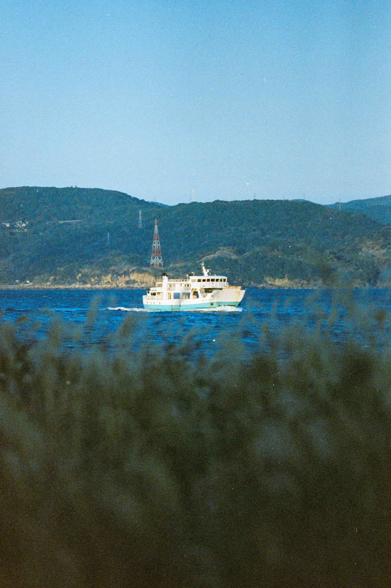 A white and teal ferry crosses the channel at Ikeshima, trailing a bright wake, with a red and white lattice tower rising above the forested hillside behind the port.