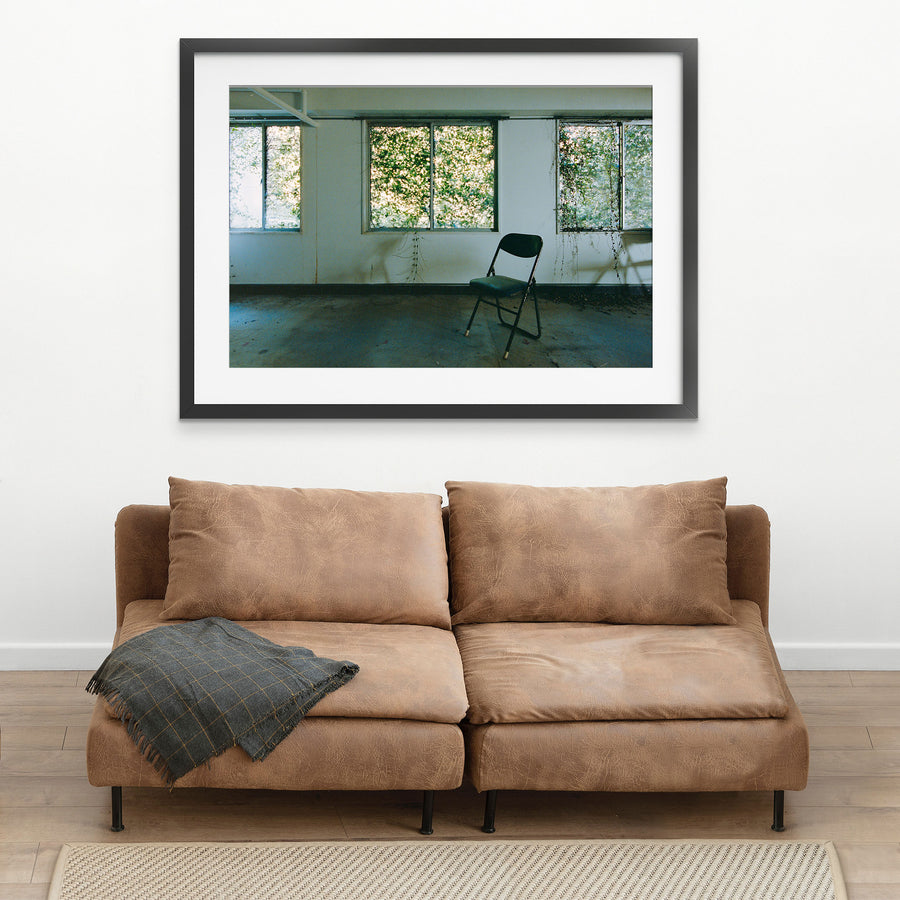 A dark green folding chair on a dusty concrete floor faces three large windows thick with green foliage, with vine tendrils trailing down the white wall beside water stains, in an office at the Ikeshima Coal Mine.