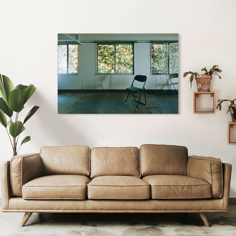 A dark green folding chair on a dusty concrete floor faces three large windows thick with green foliage, with vine tendrils trailing down the white wall beside water stains, in an office at the Ikeshima Coal Mine.
