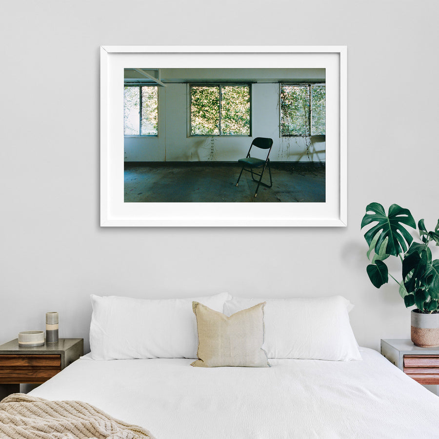 A dark green folding chair on a dusty concrete floor faces three large windows thick with green foliage, with vine tendrils trailing down the white wall beside water stains, in an office at the Ikeshima Coal Mine.