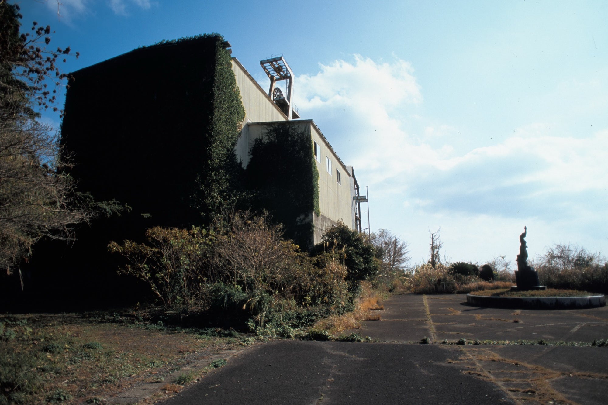 A dark statue on a circular plinth in an overgrown planter at the base of a concrete apartment block at Ikeshima, with the mine headframe visible on the roofline against a bright sky.
