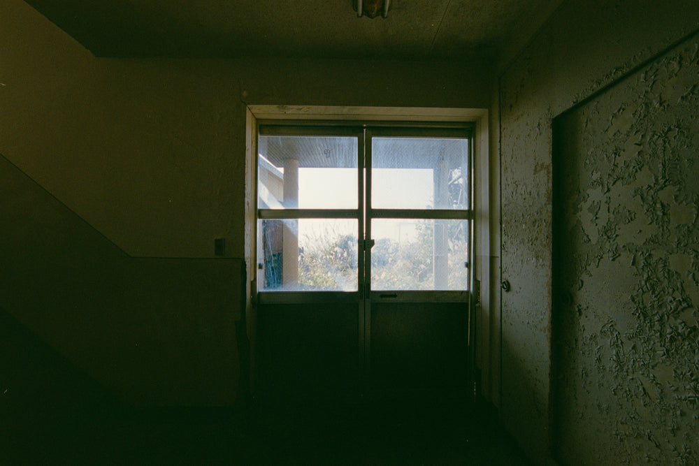 Glazed sliding doors at the end of a dark hallway in the No. 2 Shaft office at Ikeshima Coal Mine, tall grass outside the wired glass.