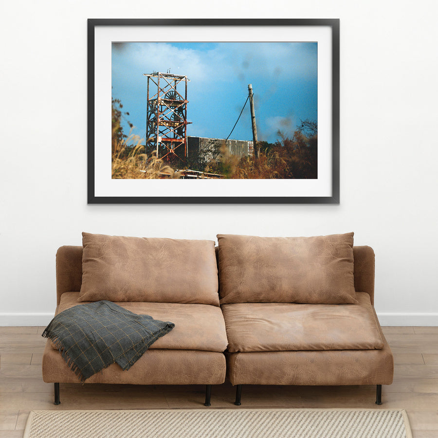 A rusting steel lattice headframe with two spoked sheave wheels silhouetted against a bright sky at Ikeshima Coal Mine, with dry grasses blurring across the foreground.